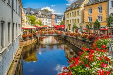 Kanal fließt durch Saarburgs Altstadt, gesäumt von roten und weißen Blumen. Bunte historische Gebäude und rote Sonnenschirme entlang des Wassers.