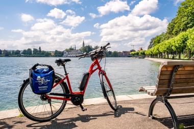 Rotes Fahrrad mit blauen Taschen mit der Aufschrift 'Radweg Reisen' neben einer Bank an einer Seepromenade. Kirchtürme und Bäume am gegenüberliegenden Ufer.