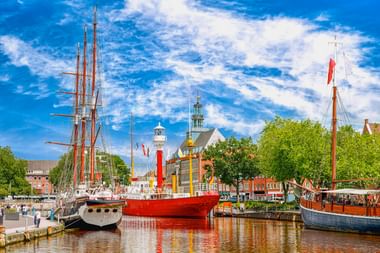Drei historische Schiffe im Emder Hafen mit einem roten Feuerschiff in der Mitte. Kirchtürme und bunte Gebäude säumen das Ufer unter blauem Himmel.