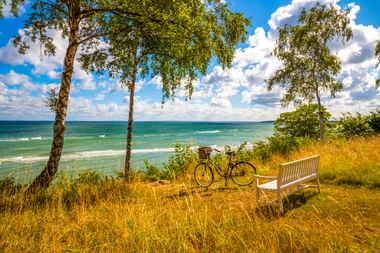 Fahrrad und weiße Bank auf grasbewachsenem Hang mit Blick aufs Meer in Munkerup. Birken rahmen die Aussicht unter blauem Himmel mit weißen Wolken.