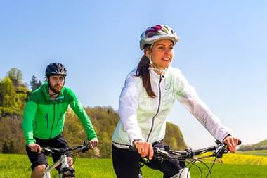 Zwei Radfahrer fahren durch grüne Landschaft am Bodensee. Frau in weißer Jacke führt, Mann in grüner Jacke folgt an sonnigem Tag.