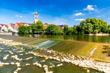 Blick auf Nürtingen am Neckar mit seichtem Wasser, felsigem Flussbett, grünen Bäumen und weißem Kirchturm über historischen Gebäuden.