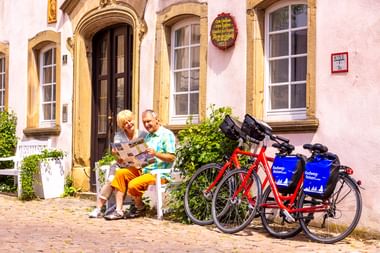 Zwei Radfahrer sitzen auf einer Bank und lesen eine Karte vor einem historischen rosa Gebäude in Bad Wimpfen. Zwei rote Tourenräder mit blauen Taschen stehen daneben.