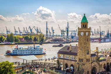 Hamburg Landungsbrücken clock tower with paddle steamer on the Elbe River. Port cranes and visitors on the pier visible under blue sky with clouds.