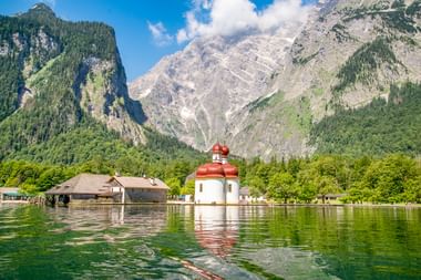St. Bartholomä Kirche mit roten Zwiebeltürmen am Königssee, umgeben von dramatischen Alpenbergen und grünen Wäldern in Bayern.