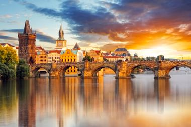 Karlsbrücke über die Moldau in Prag bei Sonnenuntergang, mit historischen Türmen und bunten Gebäuden, die sich im ruhigen Wasser spiegeln.