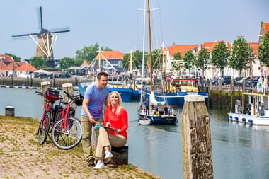 Paar mit Fahrrädern am Hafen in Zierikzee. Traditionelle Windmühle, Boote und historische Gebäude mit orangen Dächern im Hintergrund.