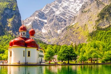 St. Bartholomä Kirche mit roten Zwiebeltürmen am Königssee-Ufer, umgeben von grüner Wiese und Bäumen mit dramatischen Alpengipfeln dahinter.