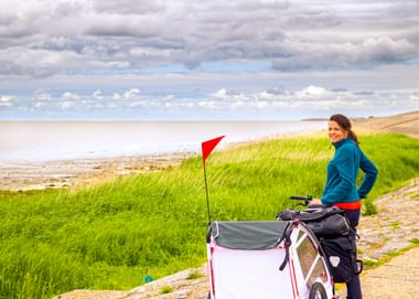 Frau in blauer Jacke mit Fahrrad und weißem Kinderanhänger mit roter Fahne auf Küstenweg in Ostfriesland. Grüne Dünen und Meer unter bewölktem Himmel.