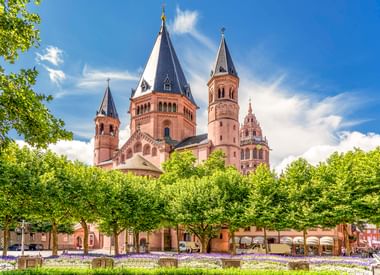 Mainz Cathedral with red sandstone towers rising above green trees. Purple flowers bloom in foreground under blue sky with white clouds.