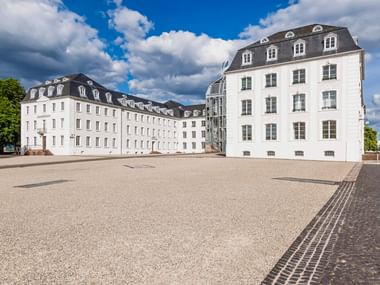 Weißes Barockschloss in Saarbrücken mit Mansarddach und Dachgauben. Großer Kiesplatz davor unter blauem Himmel mit Wolken.