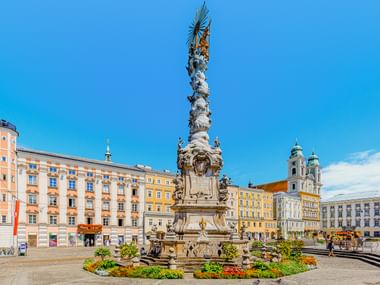 Barocke Dreifaltigkeitssäule am Linzer Hauptplatz, umgeben von farbenfrohen historischen Gebäuden und einer Kirche mit Zwillingstürmen.