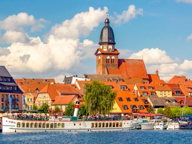 Harbor view of Waren Müritz with passenger boat, traditional buildings with orange roofs, and prominent church tower under blue sky with clouds.