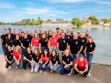Große Gruppe von Radweg-Reisen Teammitgliedern in schwarzen und roten Shirts posiert an einem See mit historischen Gebäuden und Bäumen im Hintergrund.