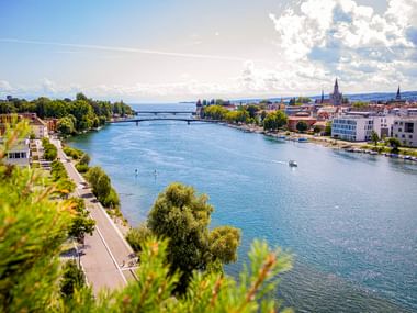 Blick auf den Seerhein in Konstanz mit Uferpromenade, Brücke und historischen Stadtgebäuden unter blauem Himmel mit weißen Wolken.