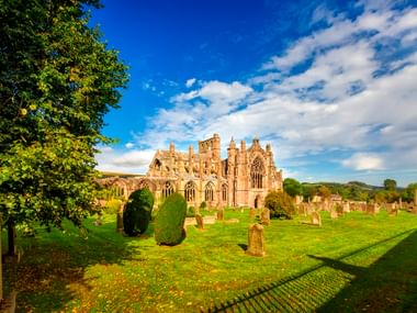 Ruinen der Melrose Abbey mit gotischer Architektur, umgeben von einem Friedhof mit Grabsteinen auf grünem Rasen unter blauem Himmel mit weißen Wolken.