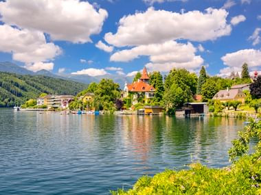 Millstätter See mit ruhigem Wasser, das Gebäude am Ufer spiegelt. Villa mit rotem Turm, grüne Bäume und bewaldete Berge unter blauem Himmel.