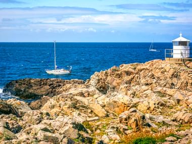 Weißer Leuchtturm auf Felsenklippen am Kullaberg mit Segelbooten auf der blauen Ostsee. Zerklüftete Küstenfelsen im Vordergrund.