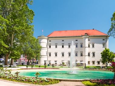 Schloss Porcia in Spittal an der Drau with white facade, red roof, and round tower. Turquoise fountain and flower beds in foreground.