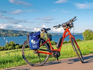Rotes E-Bike mit blauer Radweg-Reisen-Tasche auf Weg am Bodensee. Grüne Wiese, blauer See, Segelboote und Hügel unter sonnigem Himmel.