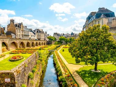 Formal gardens with water canal and colorful flowerbeds along medieval stone walls in Vannes, with historic buildings and arched bridge visible.
