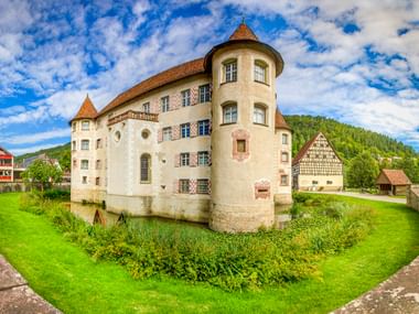 Wasserschloss Glatt in Sulz mit runden Türmen und Wassergraben, umgeben von grünem Gras. Fachwerkhäuser und bewaldete Hügel im Hintergrund.