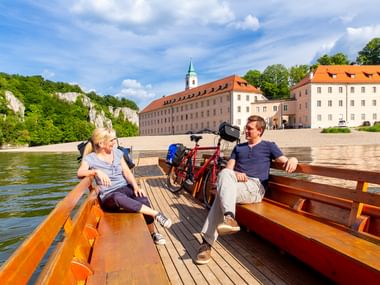Zwei Radfahrer entspannen auf einer Holzfähre mit Fahrrädern beim Überqueren der Donau nahe Kloster Weltenburg mit seinen markanten Gebäuden.