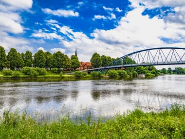 Blaue Bogenbrücke Wesertor über die Weser in Nienburg mit grünen Ufern, Bäumen und einem Kirchturm im Hintergrund.