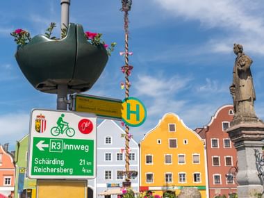 Marktplatz in Obernberg am Inn mit grünem Innradweg-Schild (R3 Innweg), bunten historischen Gebäuden, Statue und geschmücktem Maibaum unter blauem Himmel.