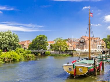 Gelb-grünes Holzboot im Hafen von Ribe mit dänischer Flagge. Historische Gebäude säumen das Ufer unter blauem Himmel mit weißen Wolken.