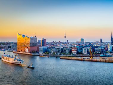 Hamburger Hafen bei Sonnenuntergang mit Elbphilharmonie, Frachtschiff und Stadtsilhouette mit Fernsehturm und Kirchtürmen entlang der Elbe.