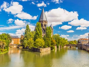 Temple Neuf Kirche mit hohem Turm auf kleiner Insel, umgeben von der Mosel in Metz. Steinbrücken verbinden die von Bäumen gesäumten Flussufer.