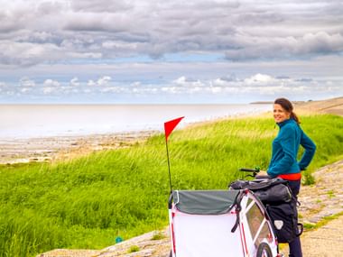Woman in blue jacket with bicycle and white child trailer with red flag on coastal path in East Frisia. Green dunes and sea under cloudy sky.