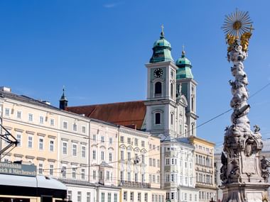 Linzer Dom mit grünen Kupferkuppeln und Zwillingstürmen neben der barocken Dreifaltigkeitssäule unter blauem Himmel.