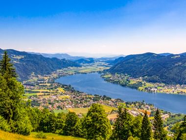 Luftaufnahme des Ossiacher Sees in einem grünen Tal mit Bergen, Dörfern und Wiesen unter blauem Himmel in Österreich.