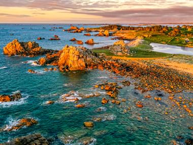 Aerial view of Gouffre de Plougrescant showing orange granite rocks scattered in turquoise water at sunset, with green coastal meadows.