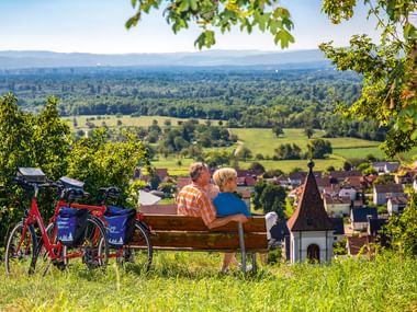 Zwei Radfahrer sitzen auf einer Holzbank neben einem roten E-Bike und blicken auf ein Dorf und grünes Tal bei Istein mit Bergen im Hintergrund.