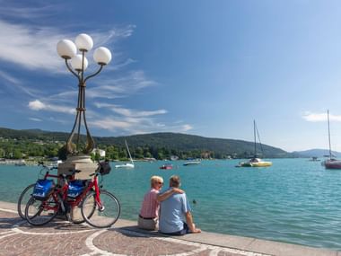 Zwei Radfahrer sitzen an der Uferpromenade in Velden am Wörthersee mit geparkten Fahrrädern. Türkisfarbener See mit Segelbooten und Bergen.