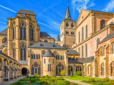 Trierer Dom mit gotischen Türmen, romanischer Architektur und formellem Gartenhof unter blauem Himmel.
