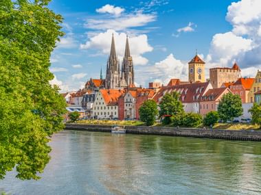 Der Regensburger Dom mit zwei Türmen erhebt sich über historische Gebäude mit roten Ziegeldächern entlang der Donau unter blauem Himmel.