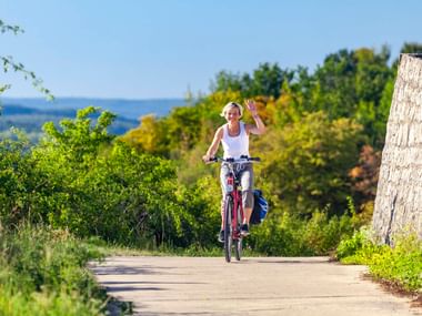 Radfahrerin winkt beim Fahren auf asphaltiertem Weg durch Zellingen Weinberge. Grüne Reben und Bäume säumen die Route unter blauem Himmel.