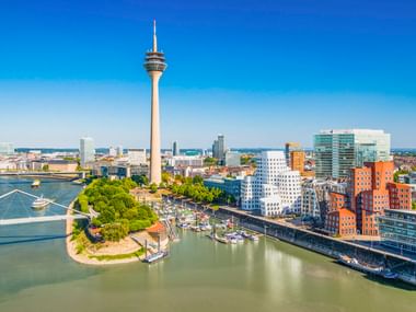 Luftaufnahme von Düsseldorf mit dem Rheinturm, Rheinhafen, modernen Gebäuden und Booten auf dem Fluss unter blauem Himmel.