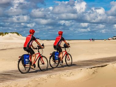 Zwei Radfahrer mit roten Helmen und Trikots fahren auf einem sandigen Strandweg in St. Peter-Ording. Sanddünen und blauer Himmel mit Wolken.