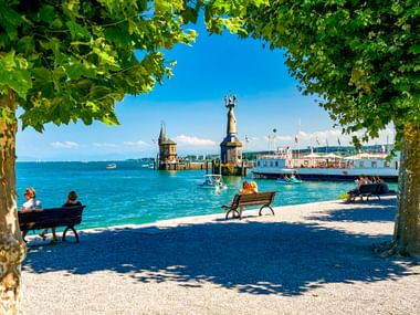Blick durch Bäume auf den Konstanzer Hafen am Bodensee mit Imperia-Statue, Leuchtturm und Booten. Menschen sitzen auf Bänken im Schatten.