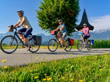 Drei Radfahrer auf einer Straße bei Rosegg mit Bildstock, großem Baum, Maisfeld und Bergkulisse unter blauem Himmel.