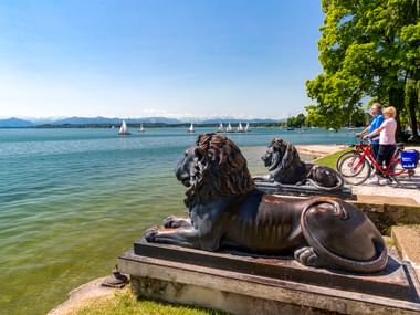 Zwei Radfahrer mit Rädern neben bronzenen Löwenstatuen am Starnberger See. Segelboote auf dem See, Berge im Hintergrund, klarer blauer Himmel.