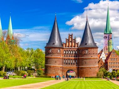 Das Holstentor, ein Backsteintor mit zwei runden Türmen, steht in einem grünen Park in Lübeck. Dahinter sind Kirchtürme und historische Gebäude sichtbar.