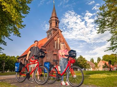 Zwei Radfahrer mit roten Tourenrädern vor der Jakobuskirche in Brunsbüttel, einer Backsteinkirche mit orangem Dach und hohem Turm, eingerahmt von Bäumen.
