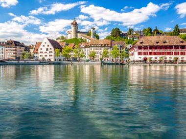 Schaffhausen Rheinpromenade mit historischen Gebäuden, die sich im ruhigen Wasser spiegeln. Turmfestung am Hang und bunte Fassaden unter blauem Himmel.