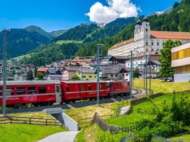 Roter Schweizer Zug auf Gleisen fährt durch Disentis mit barockem Kloster am Hang, umgeben von Alpenbergen und grünen Wiesen.